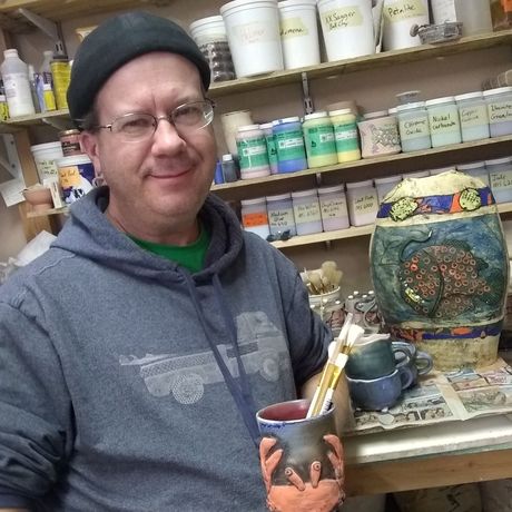 Man in a gray hoodie and black beanie holding a painted mug in an art studio with shelves of ceramic pieces