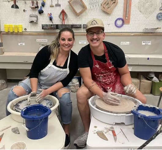 Two people smiling in a pottery studio, seated at wheels with clay and blue buckets.