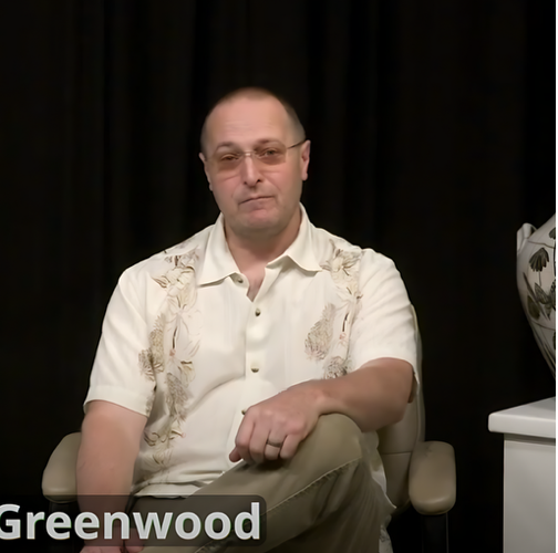 Man seated against a black backdrop, wearing a light patterned shirt, with “Greenwood” text at bottom left