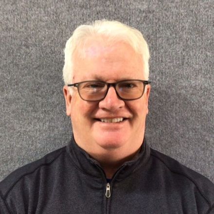 Smiling person with white hair and glasses wearing a dark zip-up jacket against a gray background