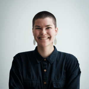 Smiling person with a buzz cut in a dark shirt against a light background