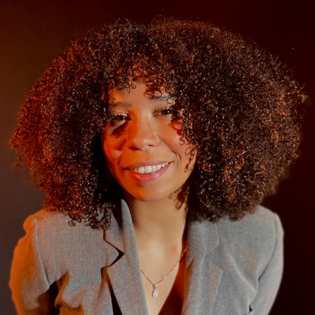 Smiling woman with curly hair in a gray blazer against a warm orange background
