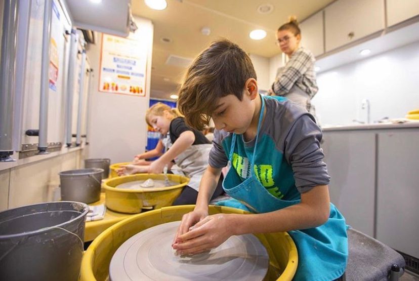 Children shaping clay on pottery wheels in the Wheel Mobile, with a teacher in the background