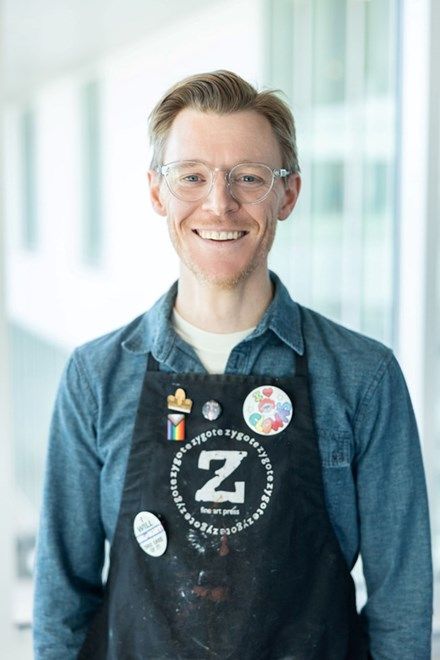 Smiling person in glasses wearing a blue shirt and black apron with pins, standing indoors.