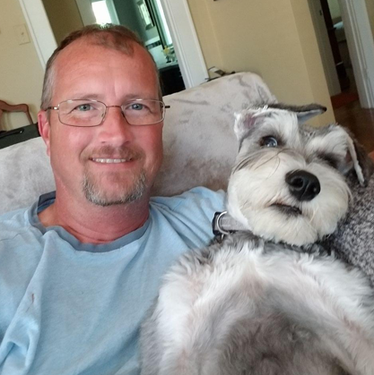 Man smiling on a couch with a small gray-and-white dog resting beside him