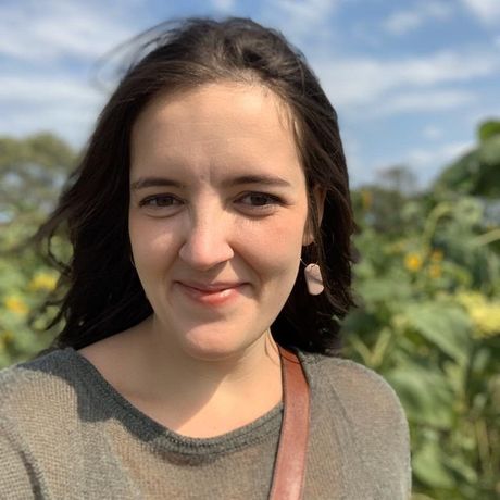 Smiling person outdoors with dark hair, wearing a gray top, in front of green plants and blue sky.