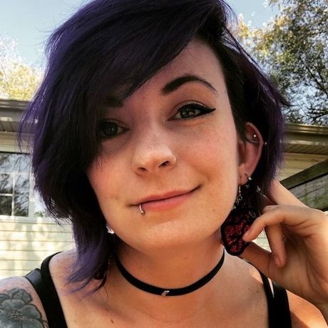 Close-up portrait of a person with dark purple hair, eyeliner, a lip piercing, and a black choker outdoors
