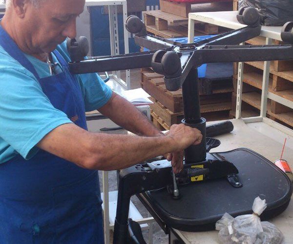 Homem de avental azul consertando uma cadeira de escritório preta em uma oficina.