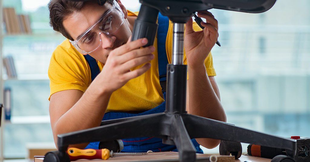 Homem usando óculos de segurança, montando uma cadeira de escritório em uma mesa, camisa amarela, close-up.