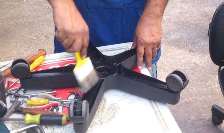 Pessoa usando uma escova para limpar a base de uma cadeira de escritório preta; ferramentas sobre a mesa.