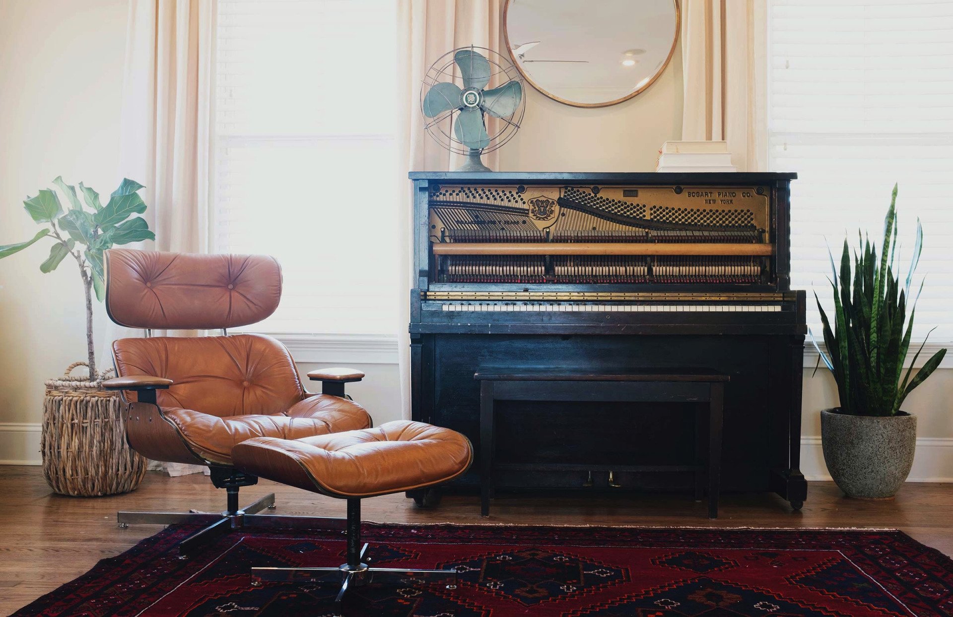 Uma sala de estar com um piano antigo, uma poltrona de couro com apoio para os pés, um tapete, plantas e um ventilador.