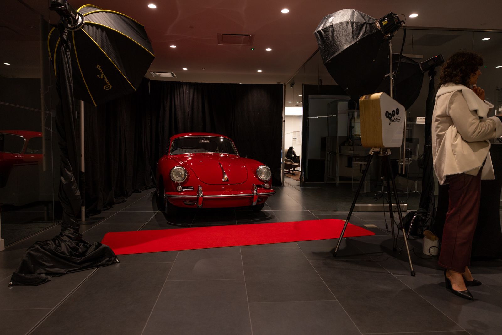 Red car on a red carpet in a photo studio with large lights and a person standing at right.