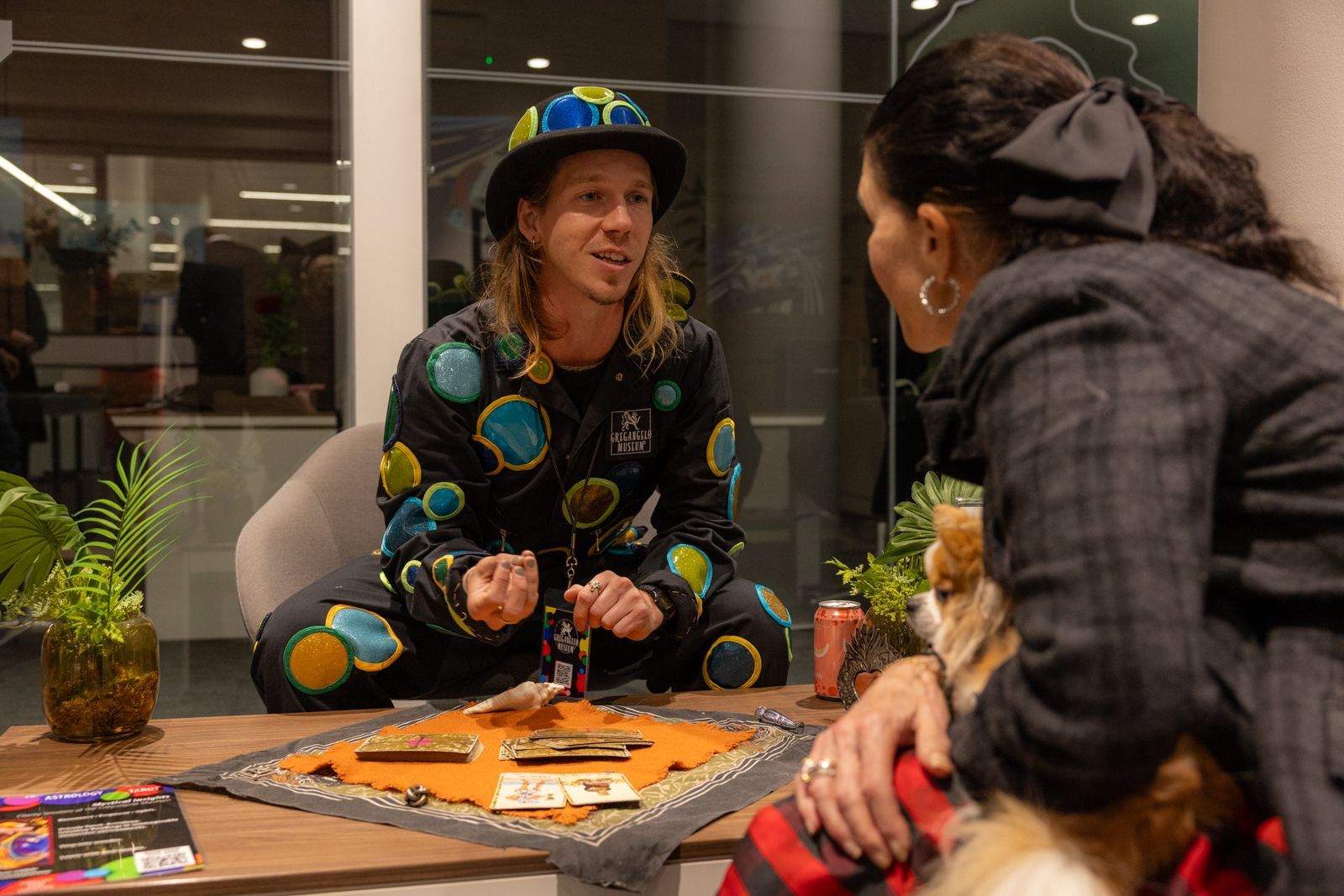 Two people talking across a table with a board game and potted plants in a glass-walled room
