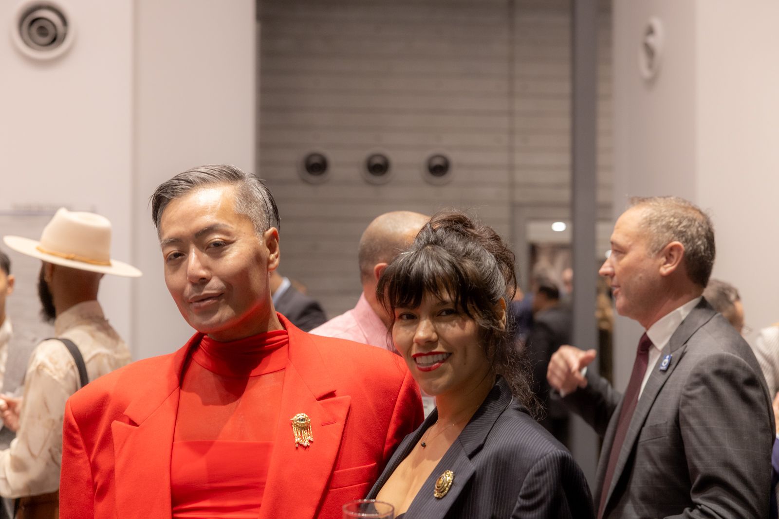 Two people in red outfits standing in a formal indoor event, surrounded by other attendees.