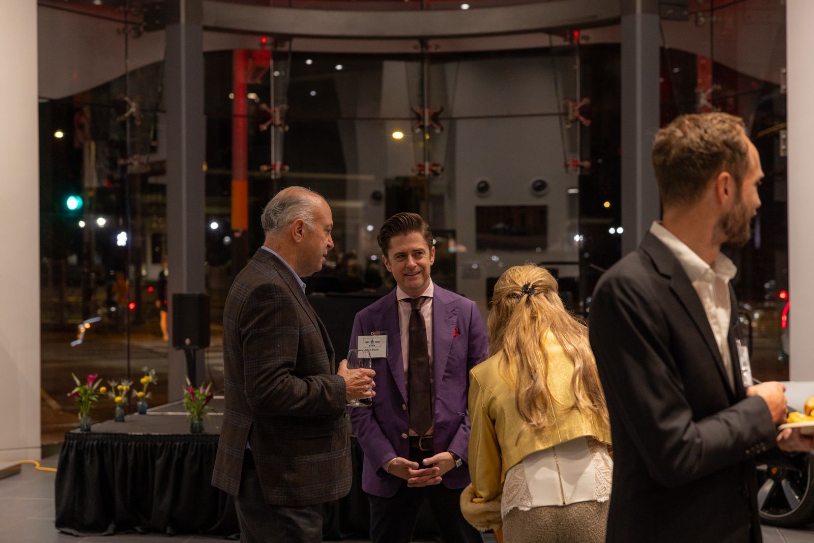 People conversing at a formal indoor event, with city lights visible through large windows.