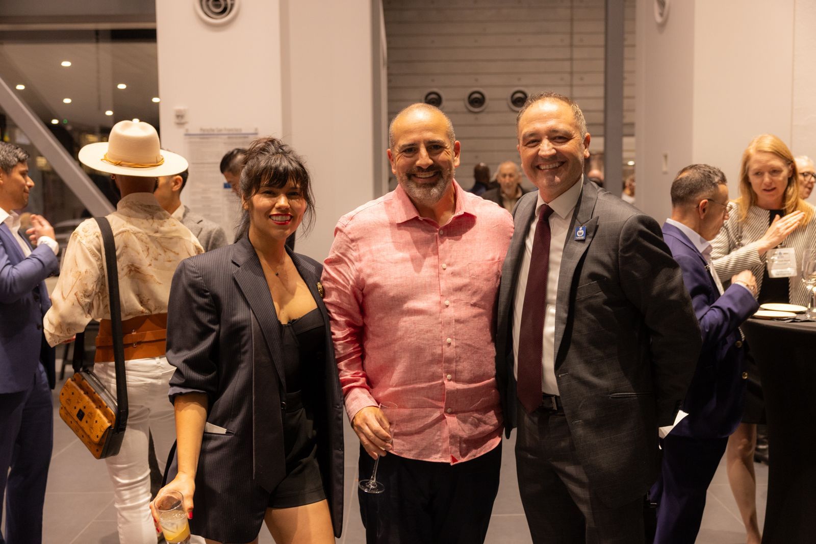 Three people posing indoors at an event, with two smiling men in dark jackets and a woman in a dark blazer.