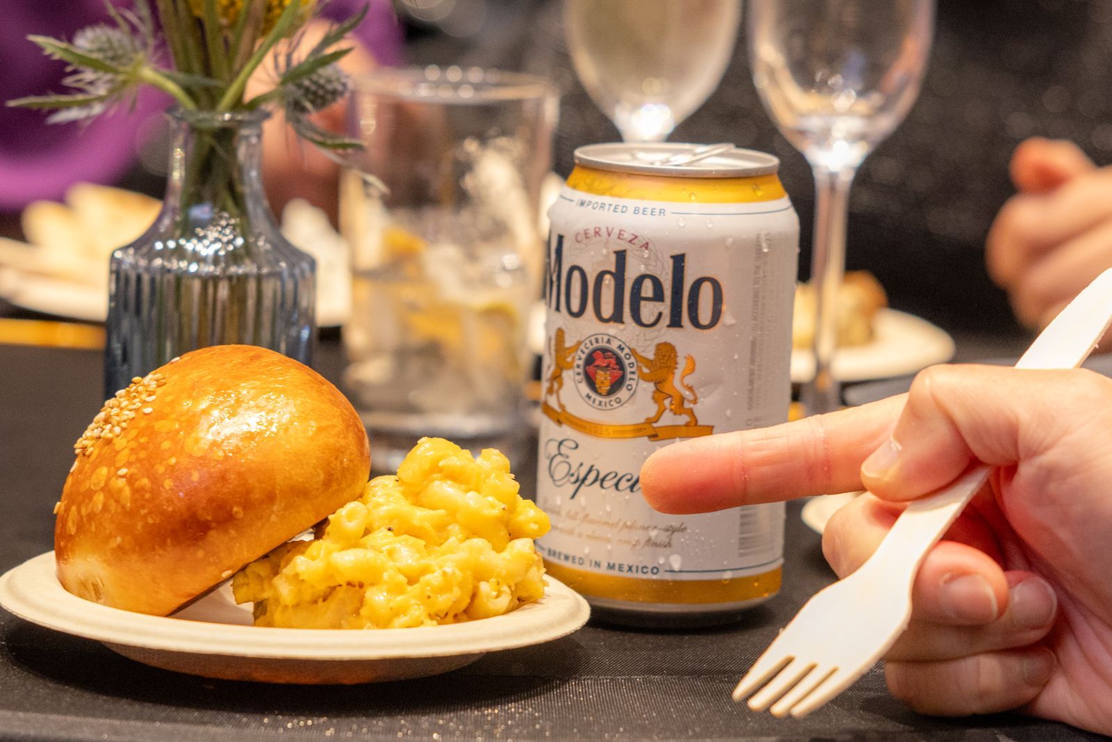Hand holding a can of Modelo beside a bun and macaroni on a table at a restaurant
