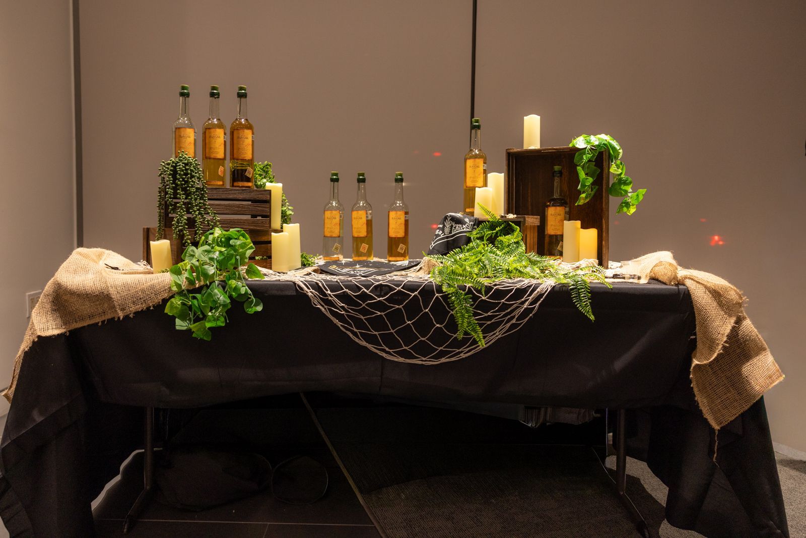 Decorated table with bottles, candles, greenery, and a black tablecloth at an indoor event