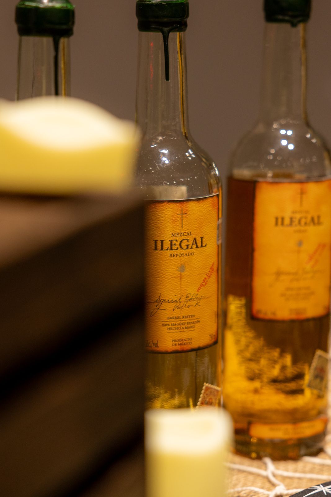 Two bottles of yellow liquor labeled “ILLEGAL” on a bar, with a blurred candle in the foreground