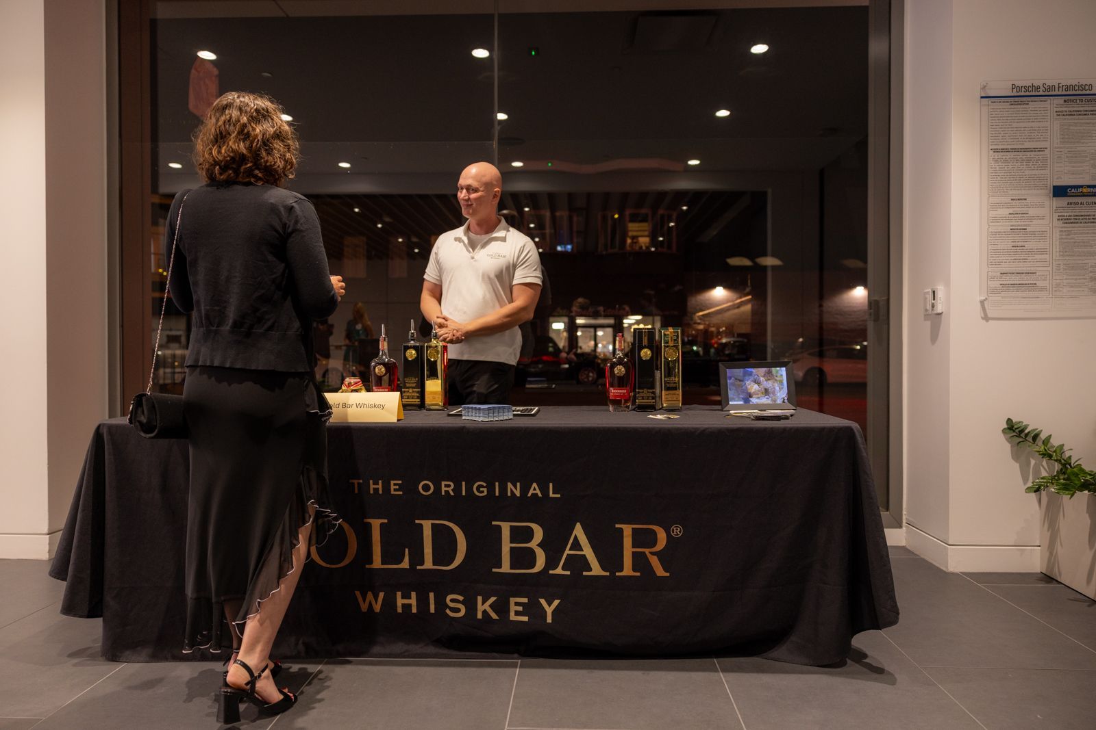 Two people at a whiskey tasting table in a lobby, with a black tablecloth reading “The Original Gold Bar Whiskey.”