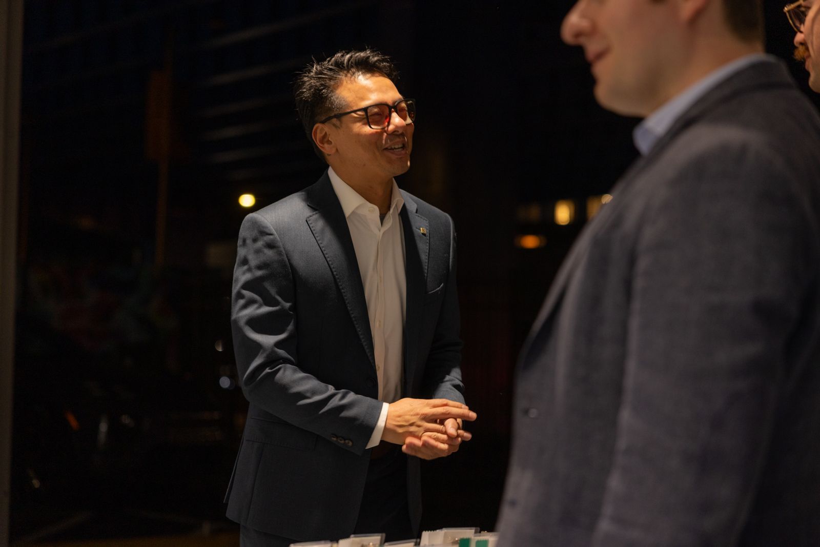 Two men in suits talking in a dimly lit room, one smiling and clasping his hands.