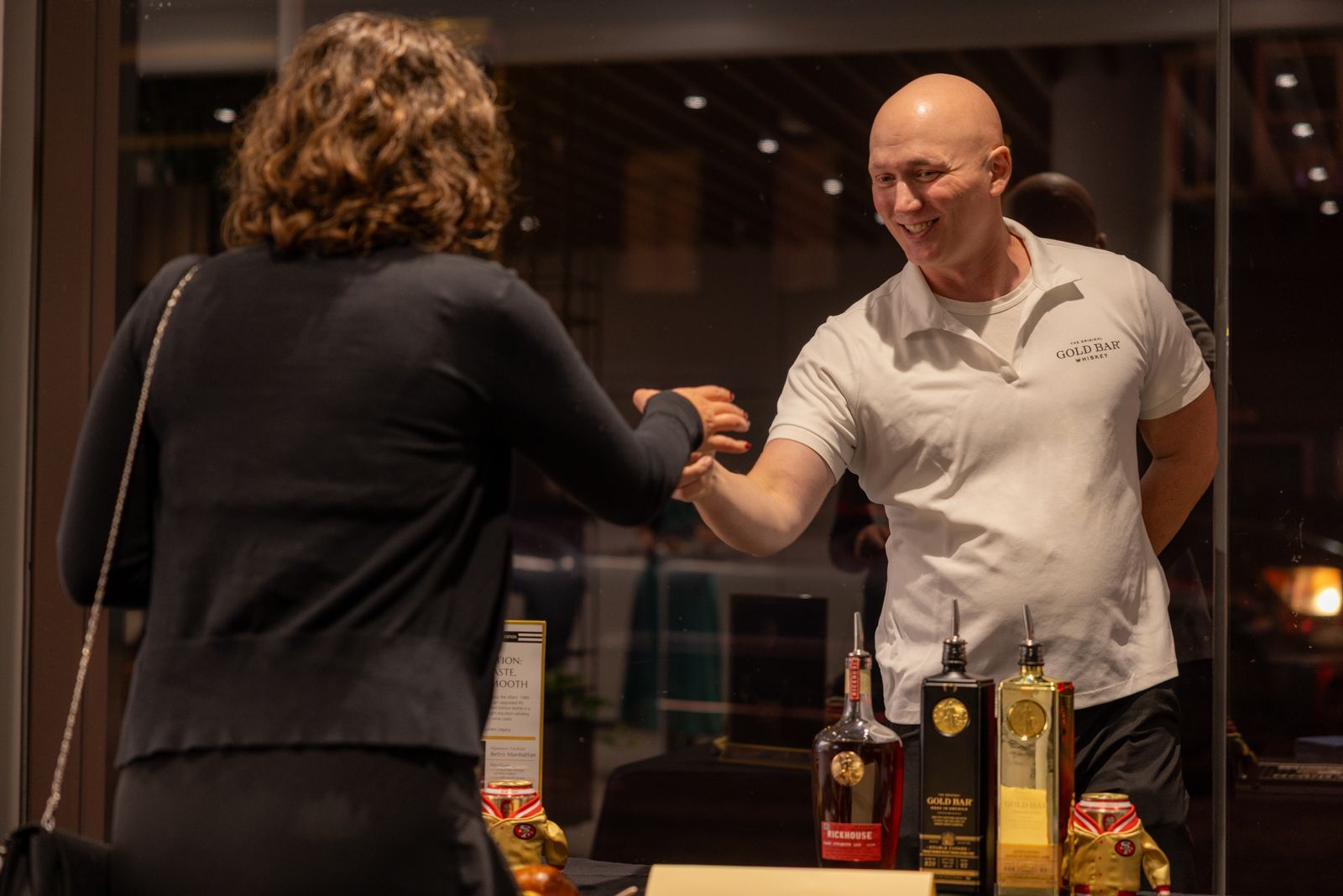 Two people shaking hands across a table with trophies and bottles in a dimly lit room