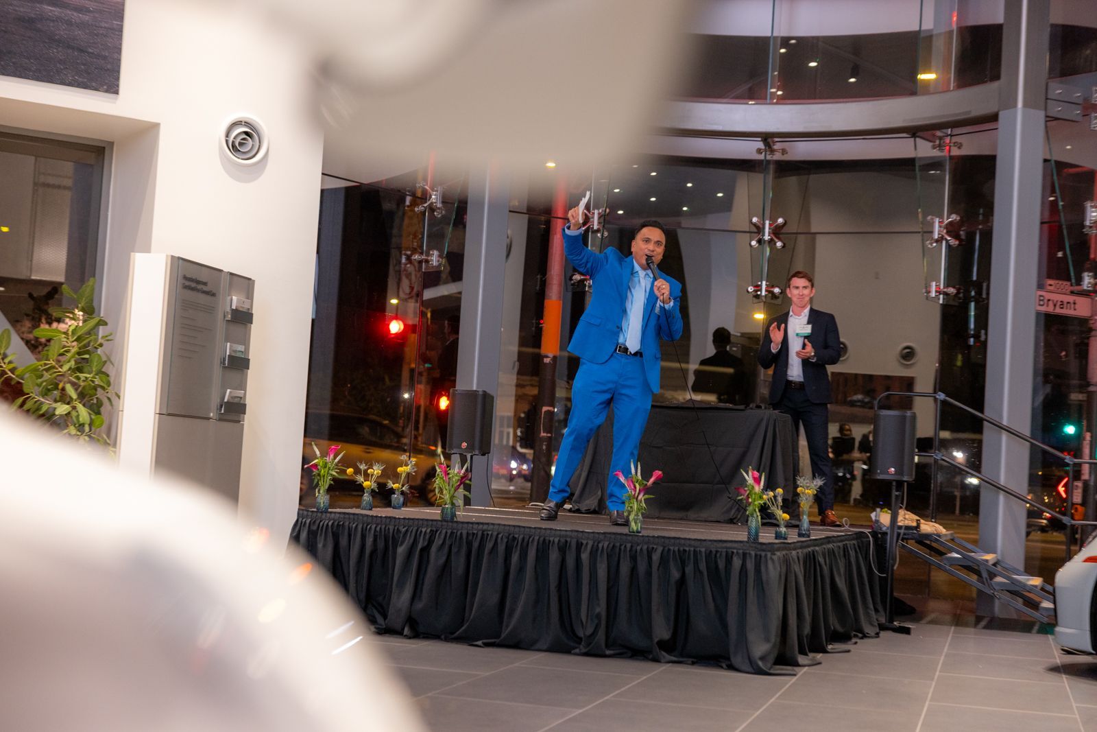 Two presenters on a small stage in a lobby, one in a blue suit and one at a podium, addressing an audience.
