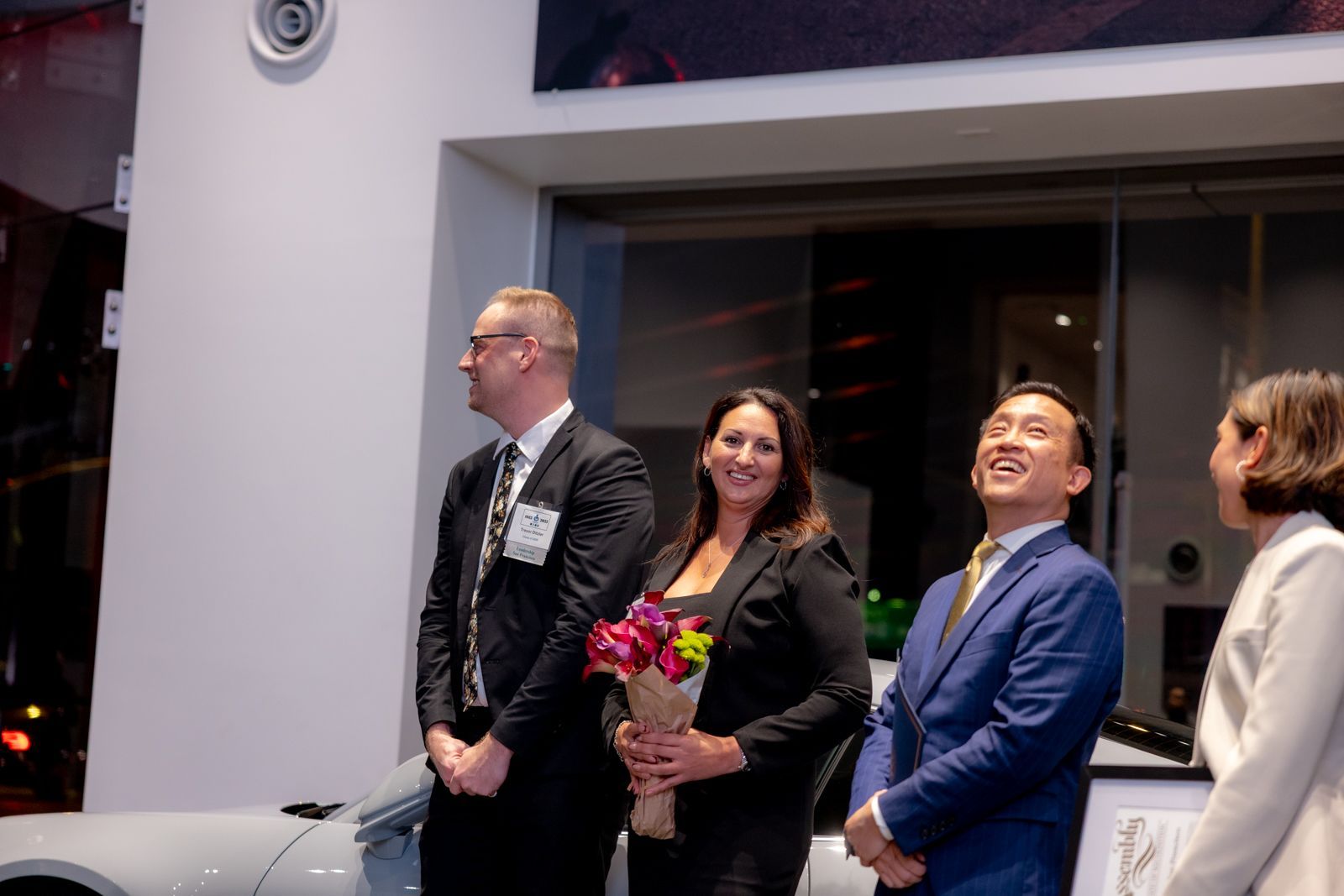 Four people in formalwear standing indoors, smiling and holding flowers at an event.