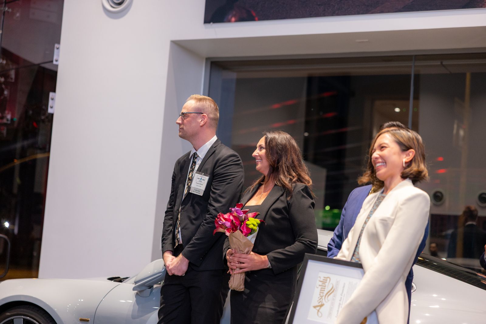 Three people stand beside a white car at an indoor event, one holding a bouquet of flowers.