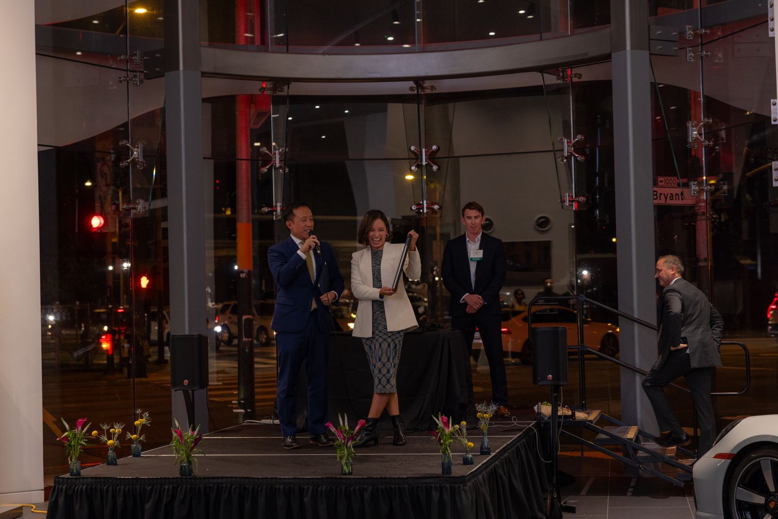 Three people on a small stage holding microphones at a nighttime event inside a glass building.