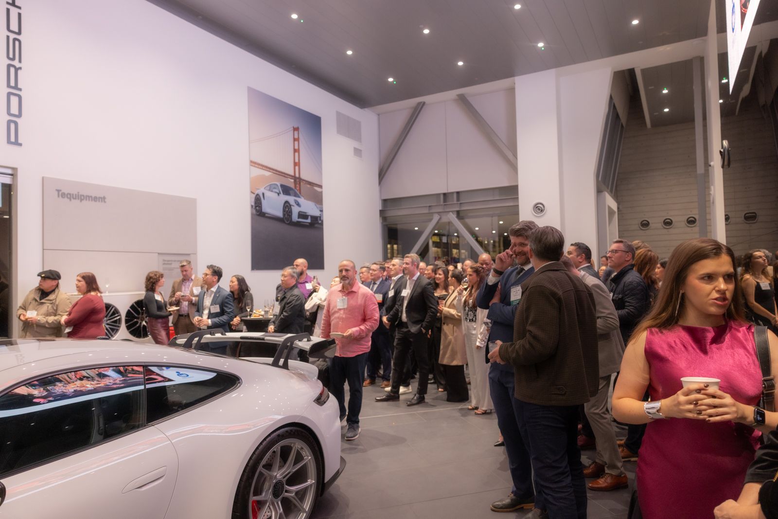 Crowd gathered inside a car showroom beside a white sports car and a large Porsche display.