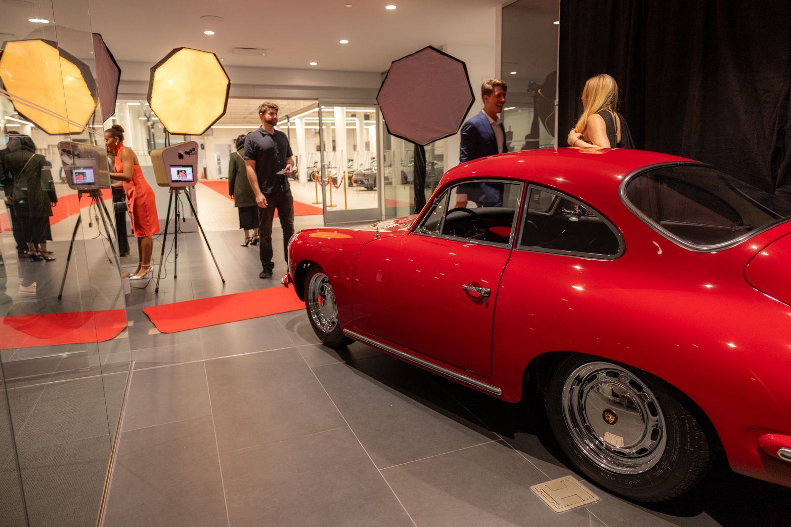 Red classic car on display in a showroom with cameras, lights, and people behind a red carpet.
