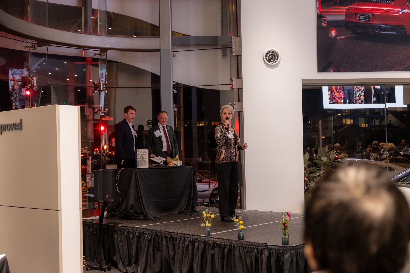 Three people stand on a small stage at an indoor event, with a large screen nearby.