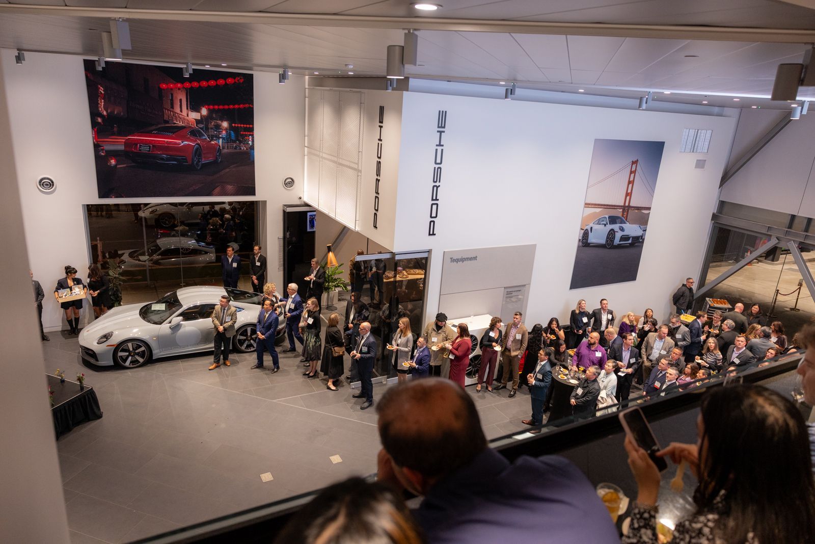 Crowded car showroom with a silver sports car on display and people gathered on a balcony and floor below