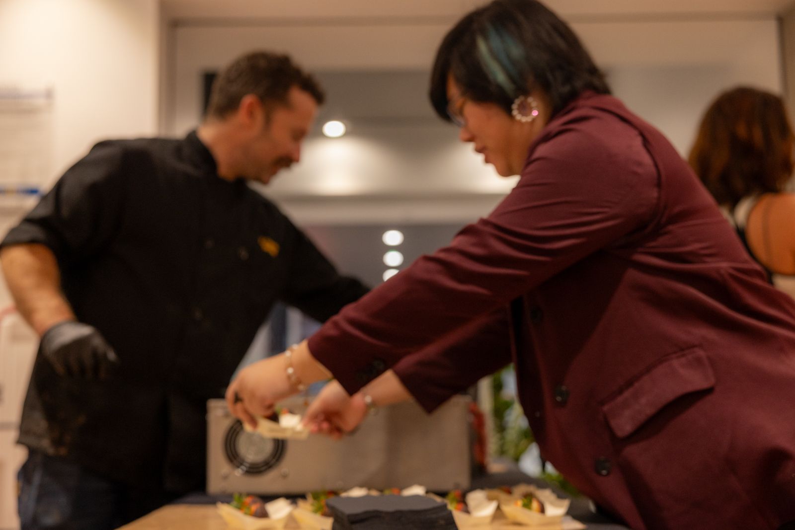 People serving food at a buffet table indoors, with one person pouring sauce onto a plate.
