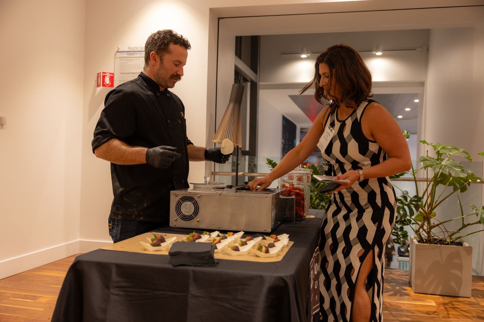 Two people serving food from a buffet table in a bright indoor hallway