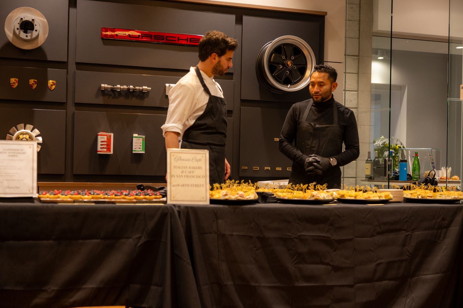 Two men behind a buffet table in a modern dining area, with trays of food and bottles on display.