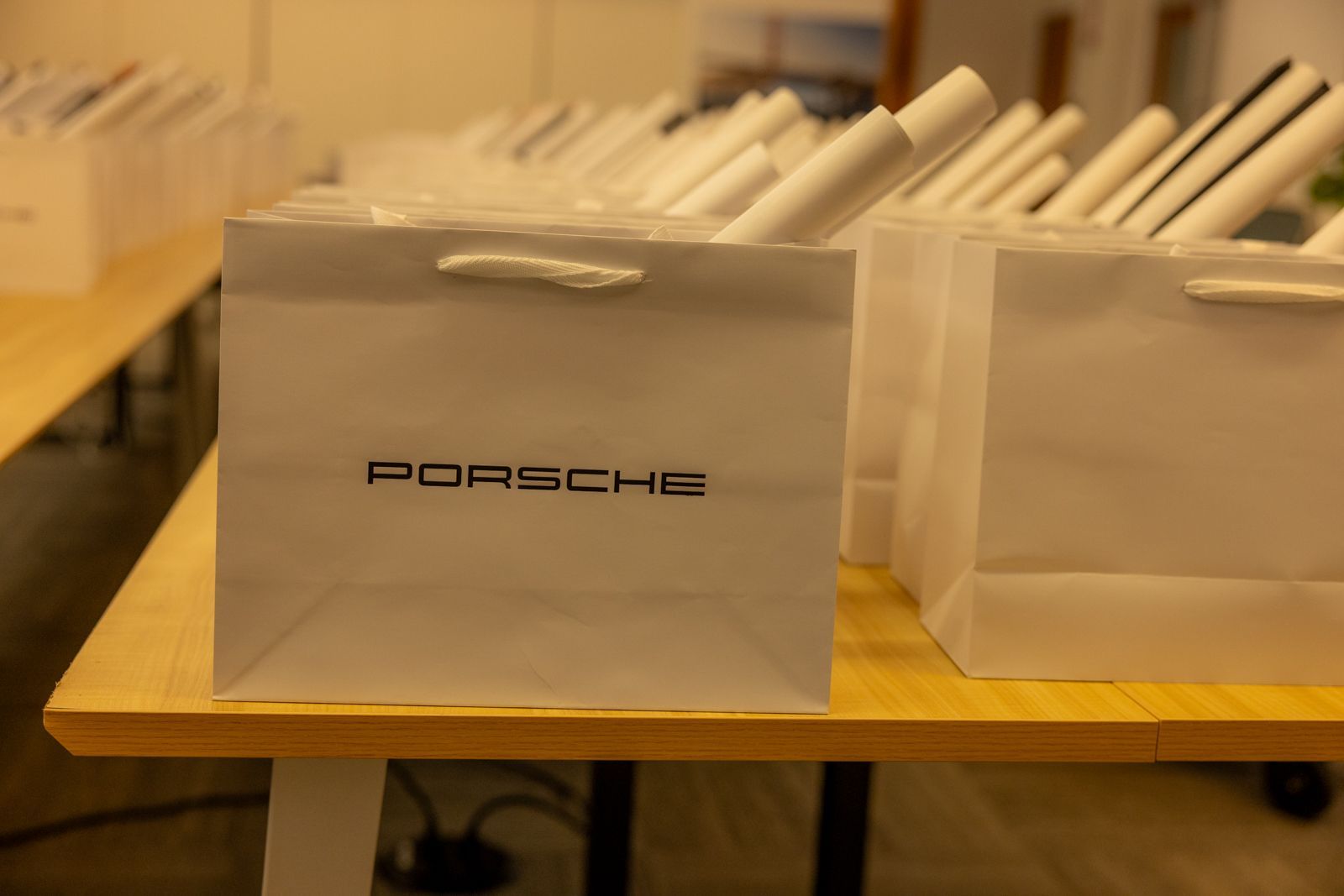 Porsche-branded gray shopping bag on a wooden table, with rolled papers in the background