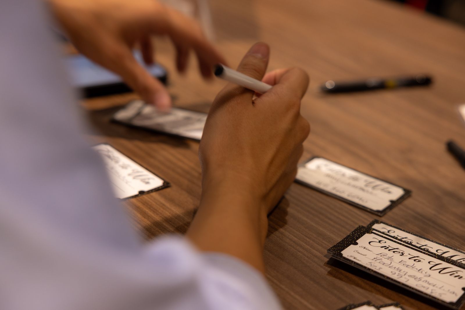 Hands writing on cards at a wooden table with pens and papers.