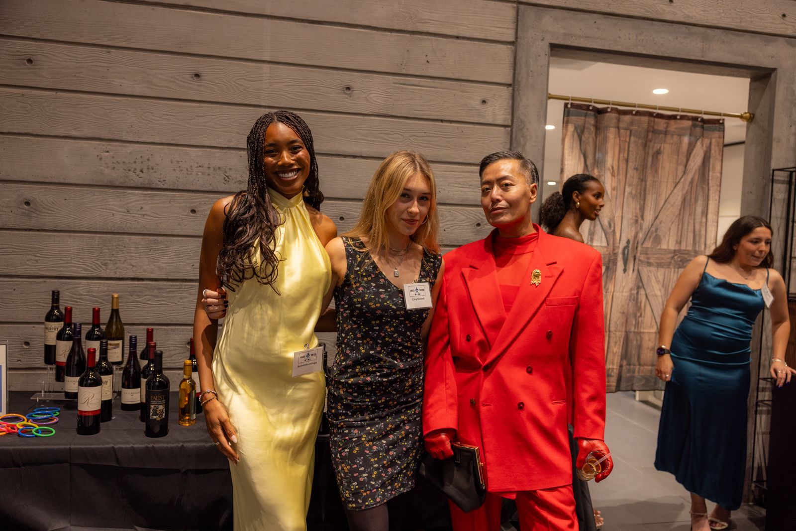 Three people posing at an indoor event, with bottles and tables in the background.