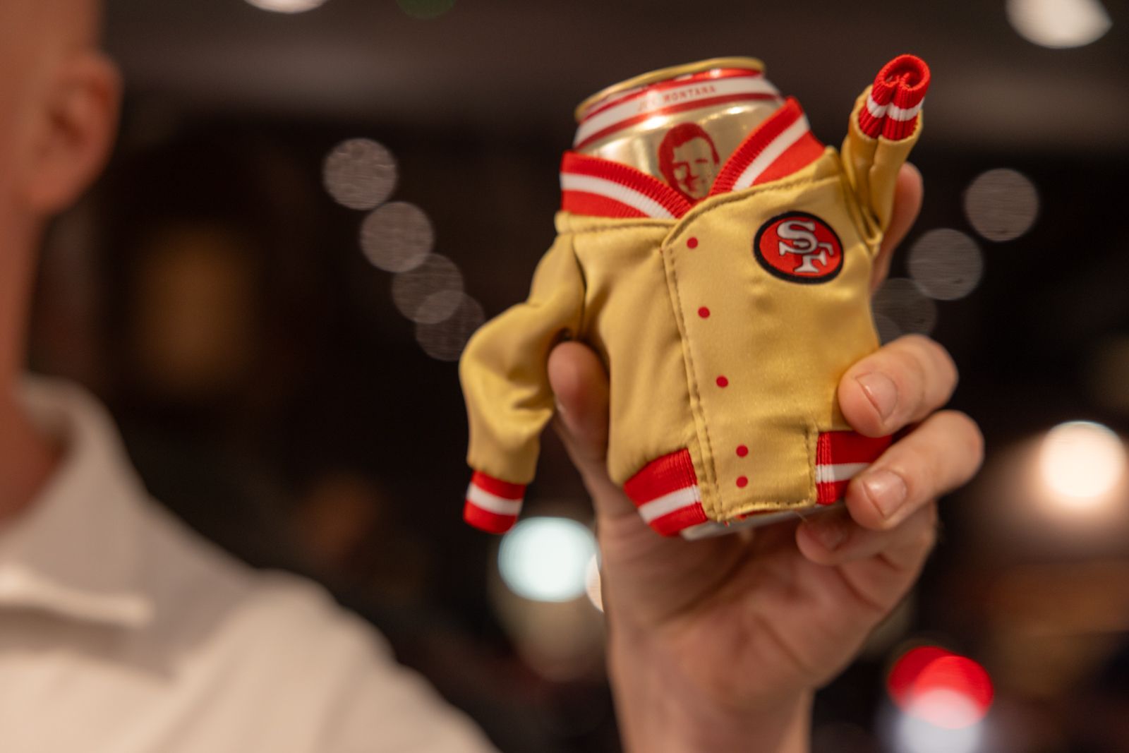 Hand holding a small red-and-gold jacket-shaped drink can in a blurred indoor setting