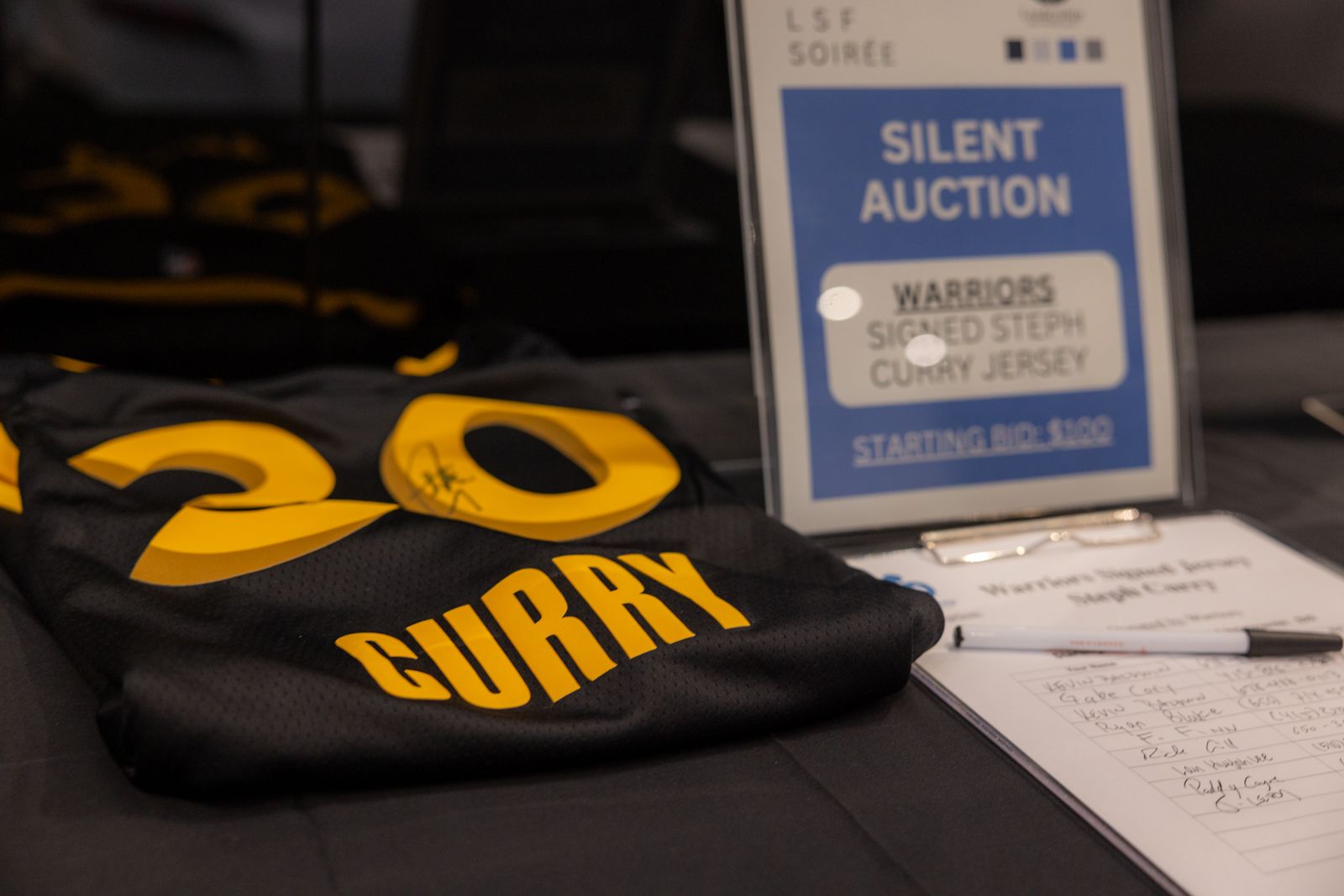 Silent auction table with black-and-yellow Curry jersey and bidding sign on display