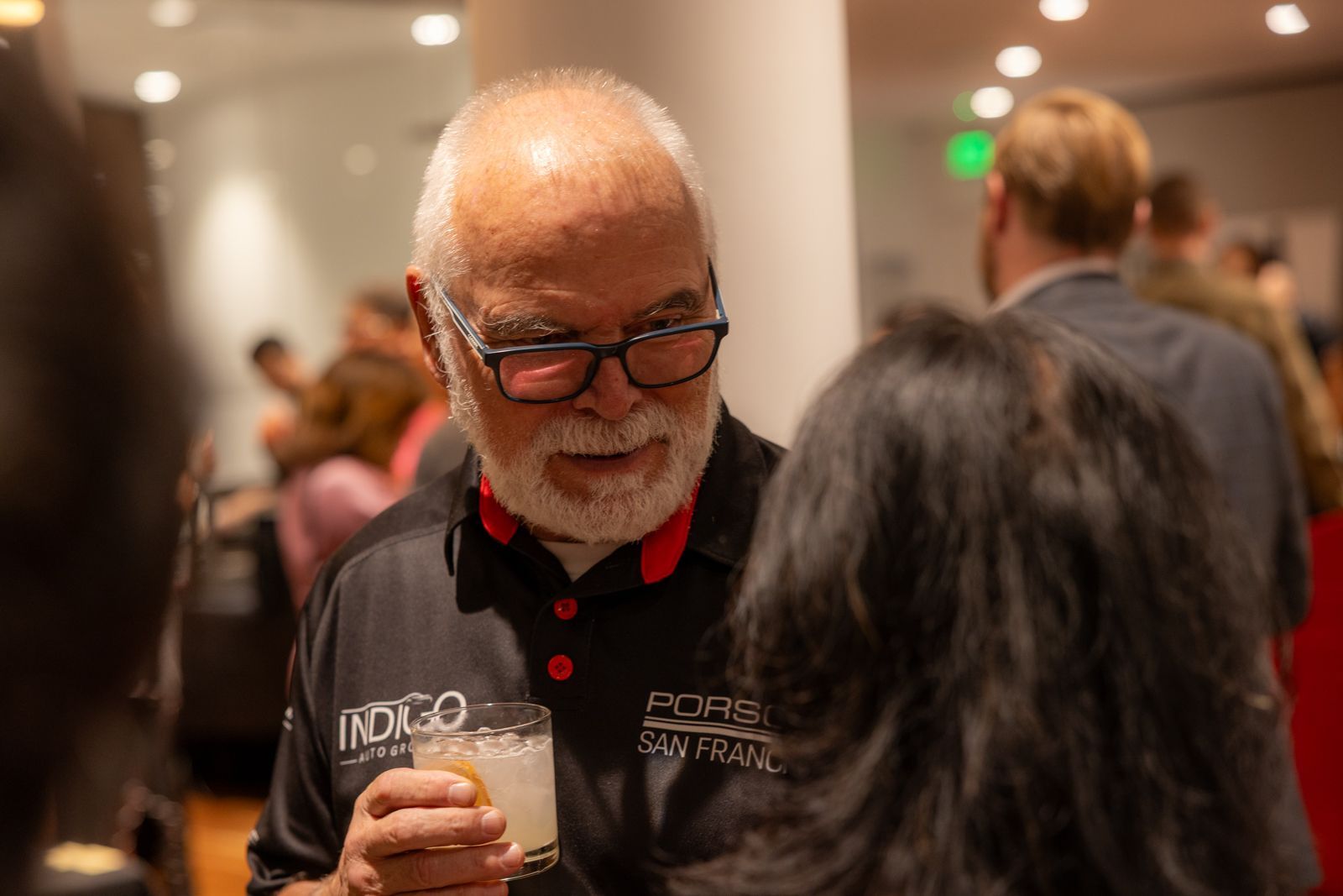Man with glasses holding a drink at a crowded indoor event, speaking to someone in the foreground
