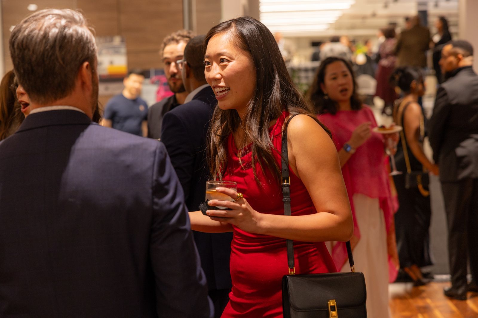 People mingling at a formal indoor event, with a woman in a red dress speaking in the foreground.