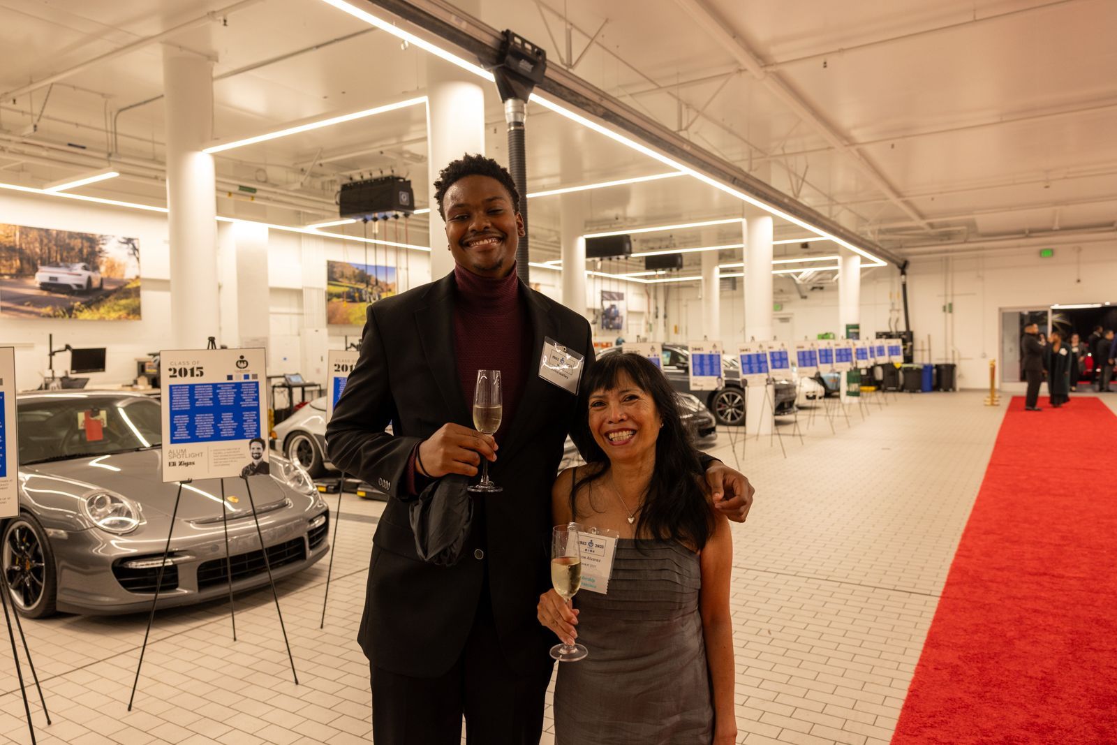 Two people holding drinks at a bright indoor event, with a silver sports car and red carpet nearby