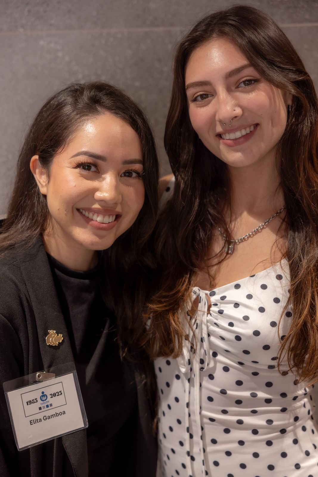Two smiling people pose indoors, one wearing a black top and name tag, the other in a white polka-dot dress.
