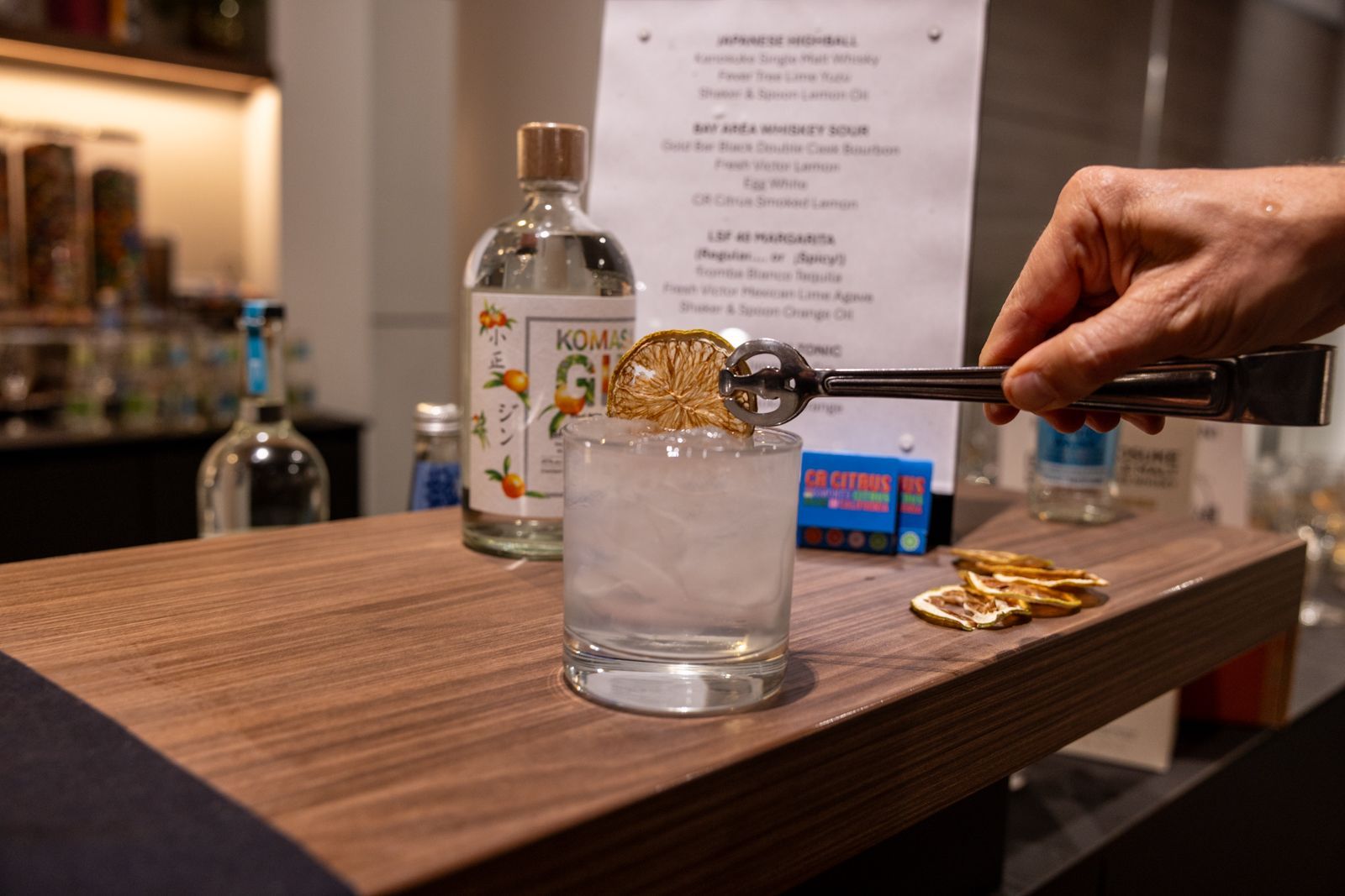 Hand pouring clear liquor into a glass with ice on a bar counter, bottles in the background