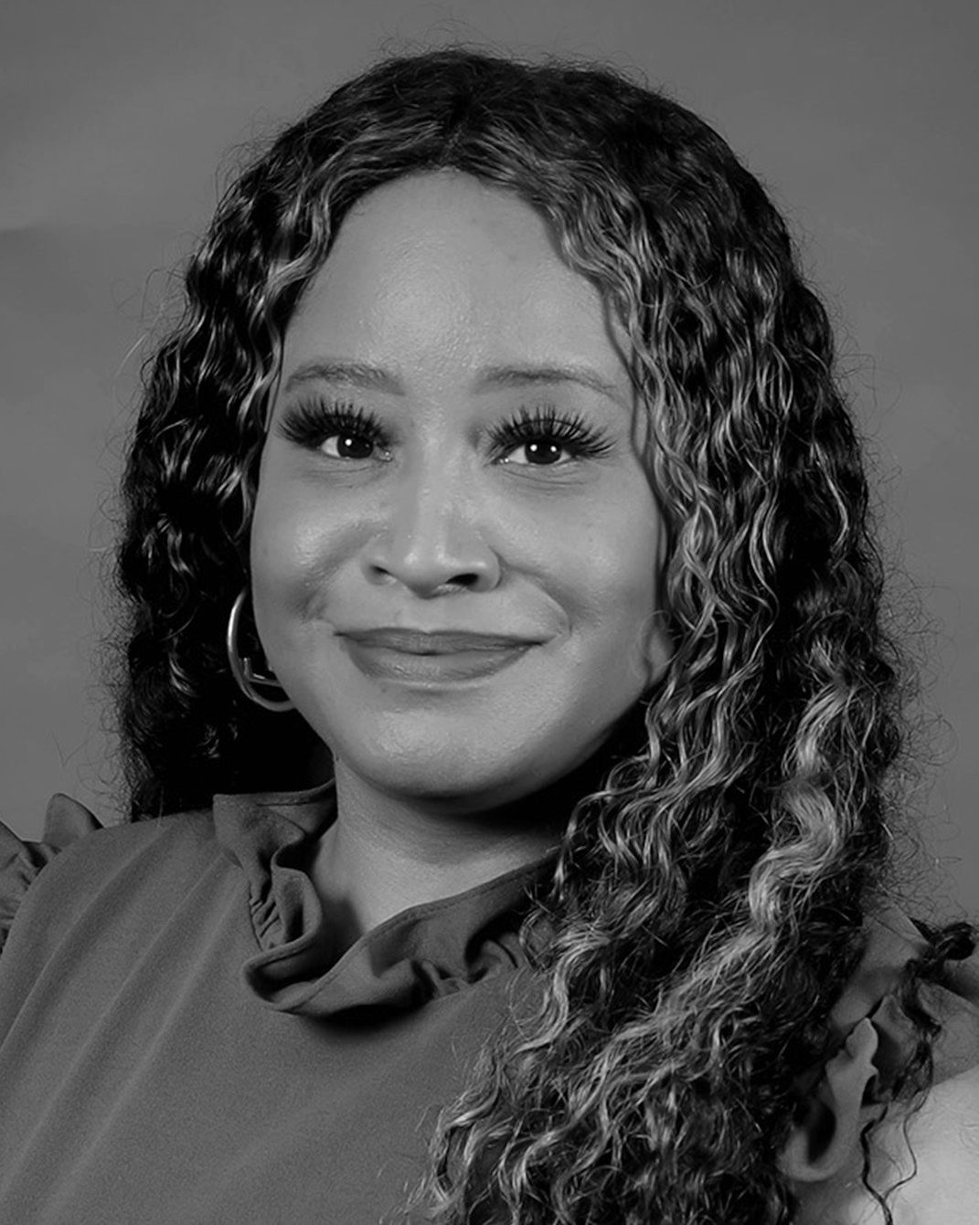 Studio portrait of a smiling person with curly hair, wearing a dark top against a gray background