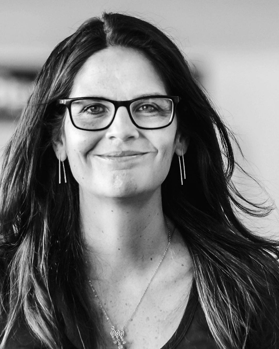 Portrait of a smiling person with glasses and long dark hair, wearing earrings and a necklace