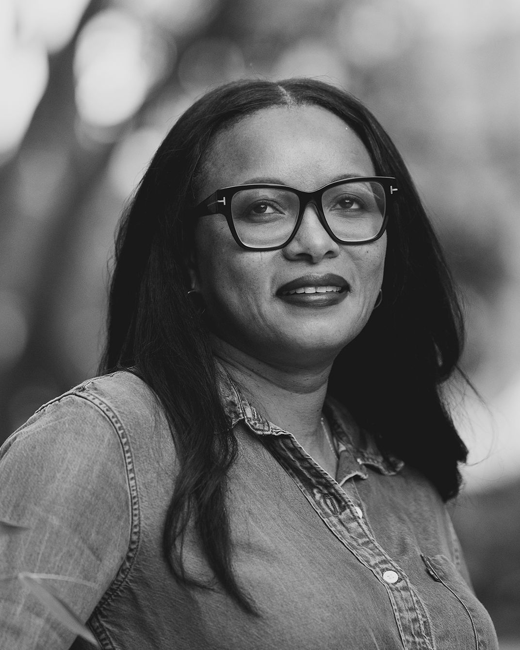 Portrait of a person with glasses and a denim shirt, smiling against a blurred outdoor background
